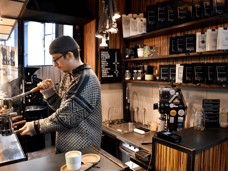 Barista připravující kávu u kávovaru a poličky plné kávových balíčků.